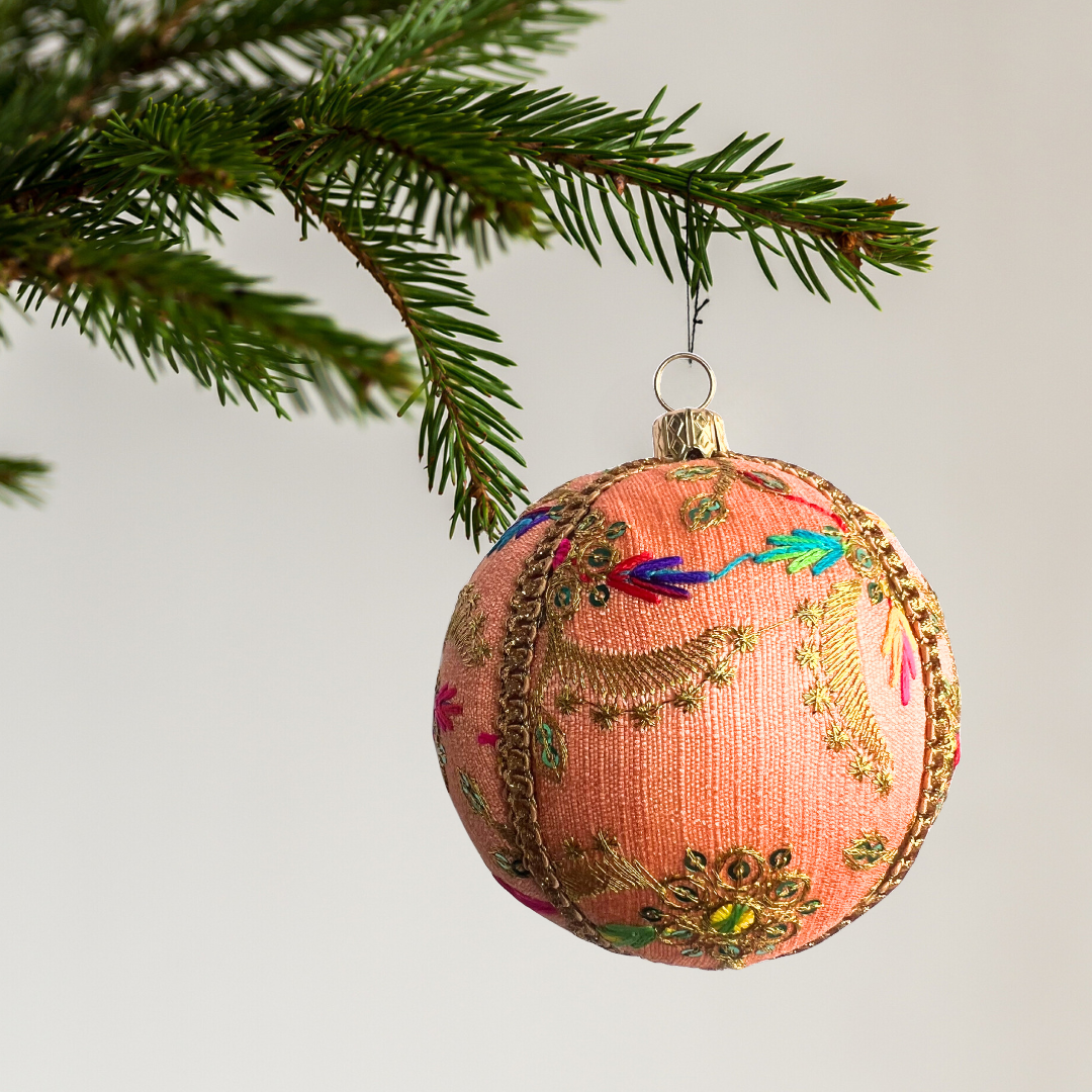 Single Aasha Bauble hanging delicately from a fir branch. The handcrafted bauble features peach fabric adorned with vibrant embroidery and gold detailing, captured against a soft white background for a clean, elegant look.